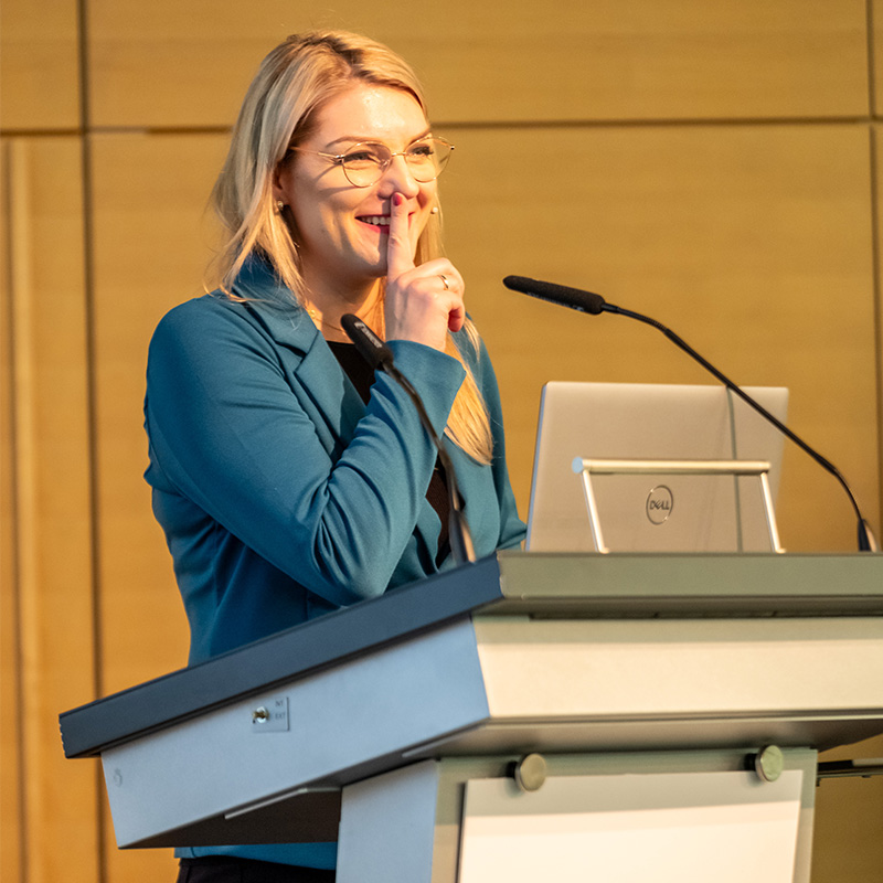 Female speaker standing behind a podium smiling, finger to lips at Petfood Forum Europe