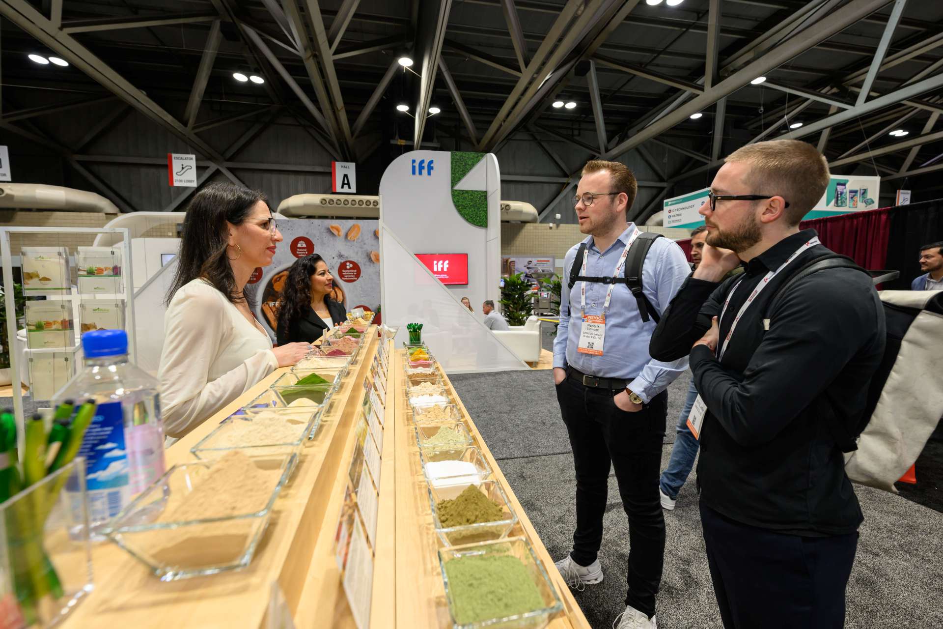 Two male attendees talk to two female exhibitors around a supplement display