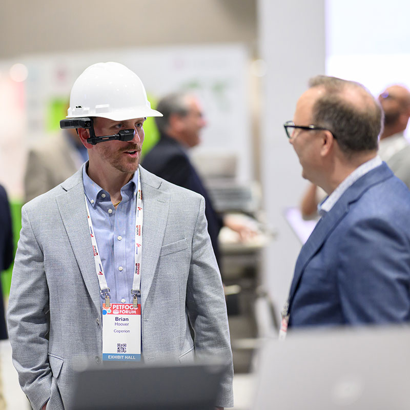 Male exhibitor wearing a hard hat from Coperion talks to an attending at Petfood Forum
